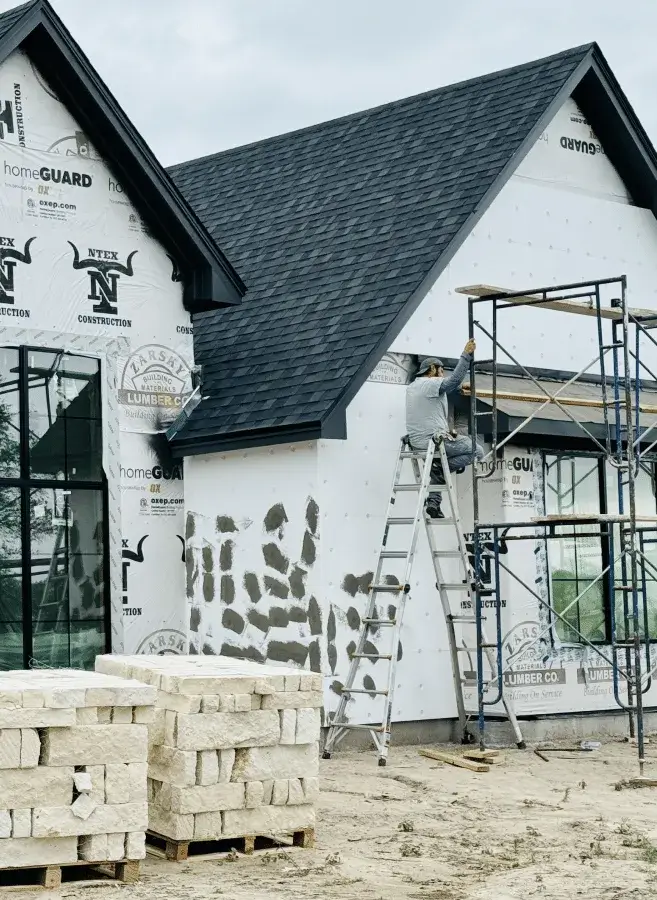Masonry worker on a ladder installing stone veneer on a house exterior; scaffolding and stacked limestone blocks sit in the foreground.