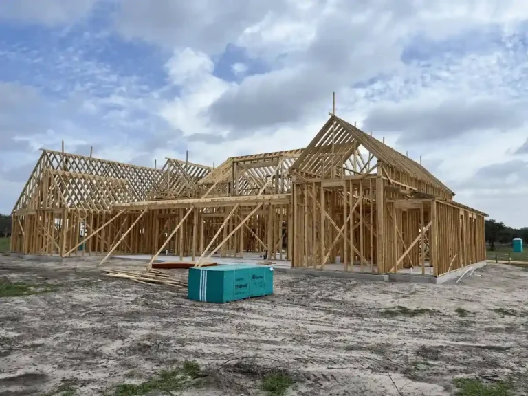 Wide view of a new home’s exposed timber frame and roof trusses rising from a concrete slab, set against a partly cloudy sky.