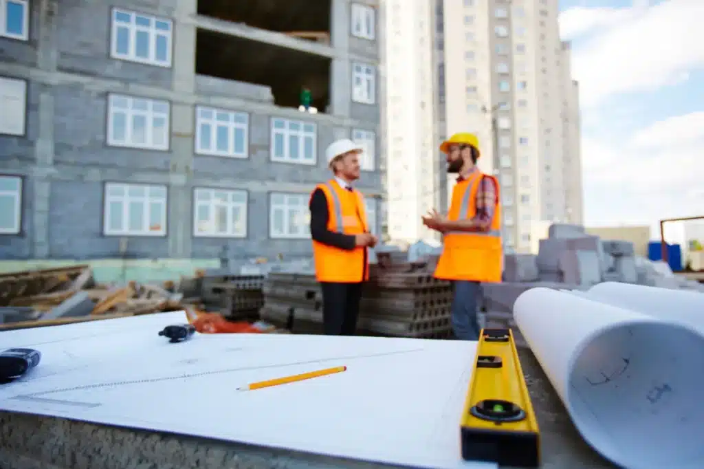 Two construction workers in hard hats and orange safety vests discussing building plans at an active construction site